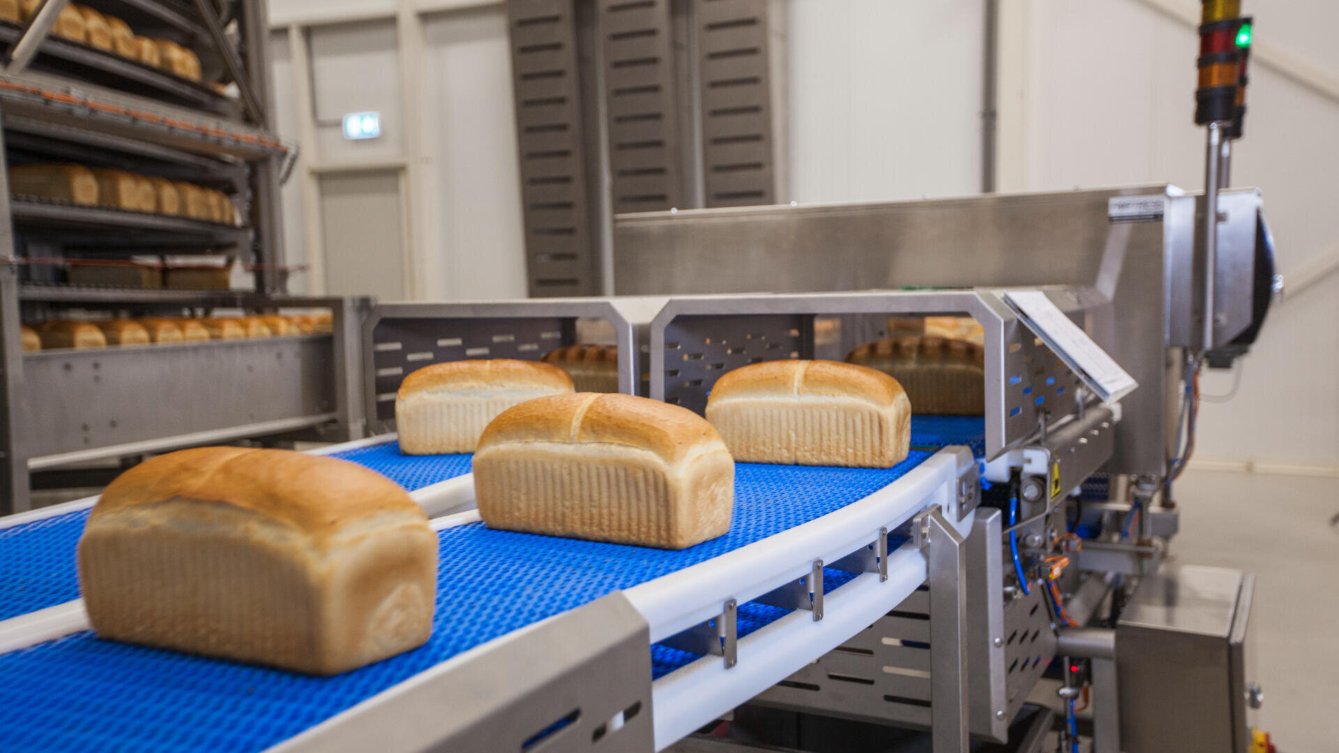 Four loaves of bread move along a blue conveyor belt in a modern bakery facility, with shelves of bread visible in the background and industrial baking equipment surrounding the area.
