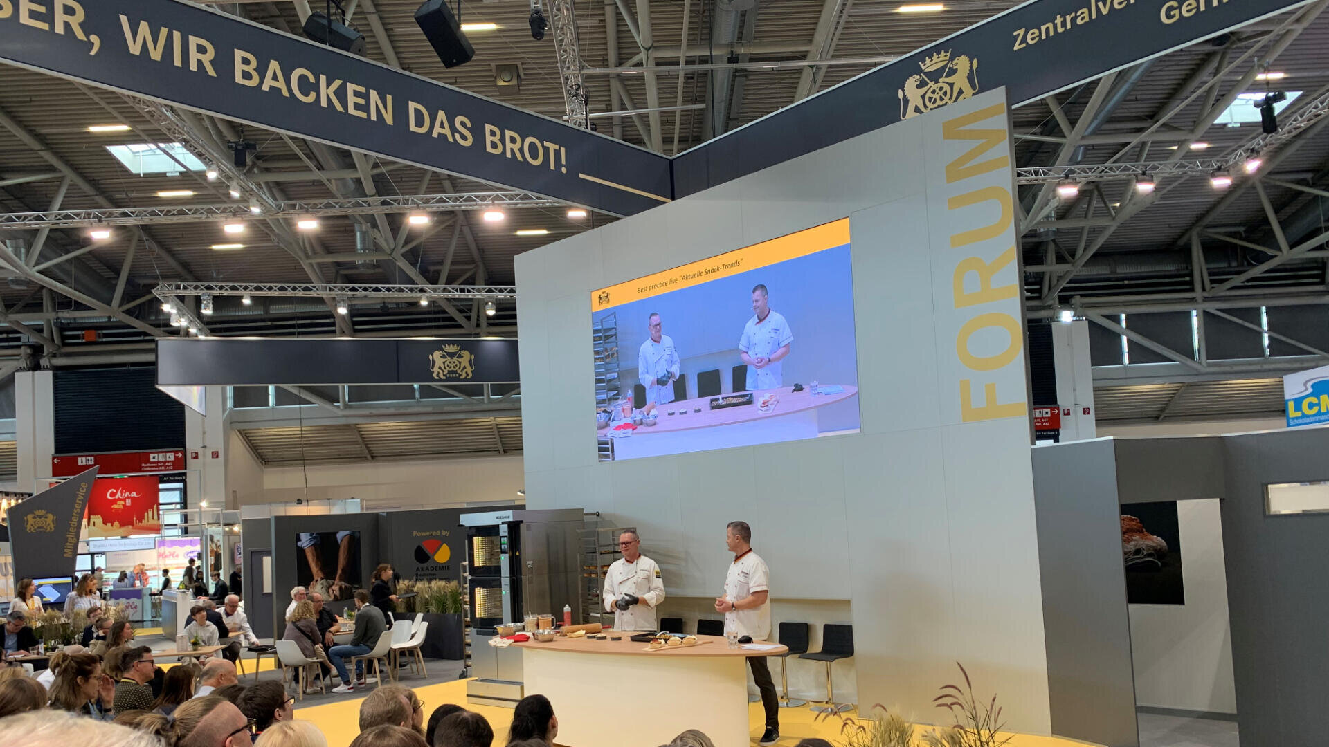 Zwei Köche demonstrieren auf einer Messebühne Backtechniken. Auf einer großen Leinwand werden ihre Aktionen übertragen. Das Publikum schaut zu, und darüber steht auf einem Banner: „Wir backen das Brot!“. Die Kulisse ist modern und gut beleuchtet.