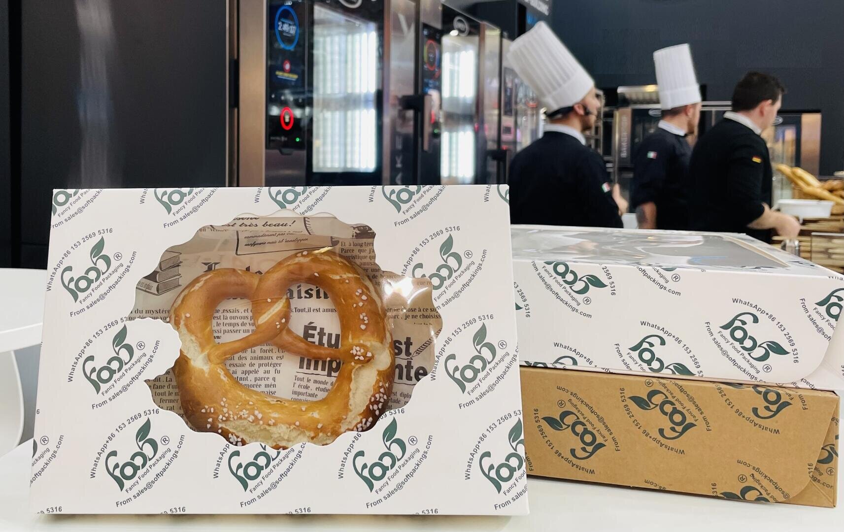 A boxed pretzel in branded packaging sits on a counter in a bakery, with bakers in chef hats working in the background near ovens.