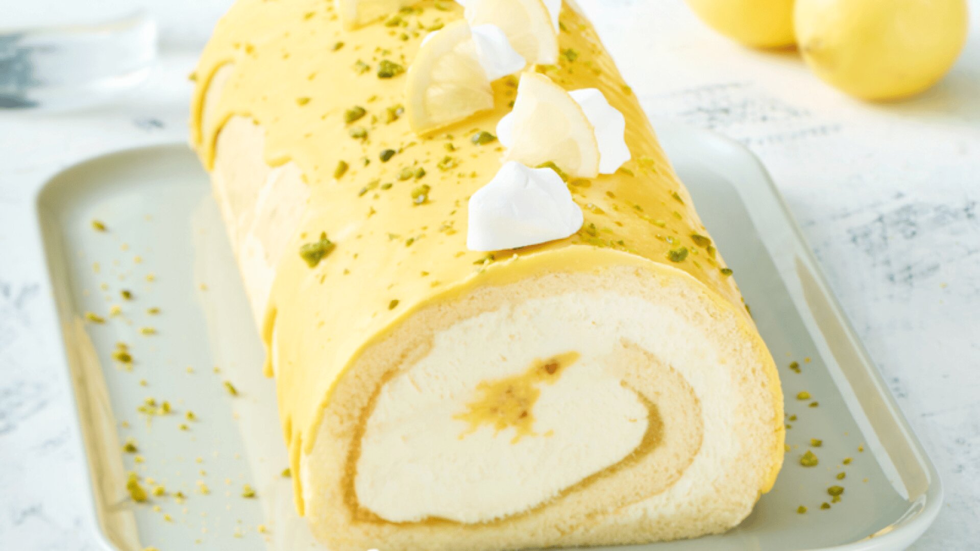 A lemon Swiss roll cake with creamy filling, topped with yellow icing, meringue pieces, and chopped pistachios, sits on a white rectangular plate. Two whole lemons are in the blurred background.