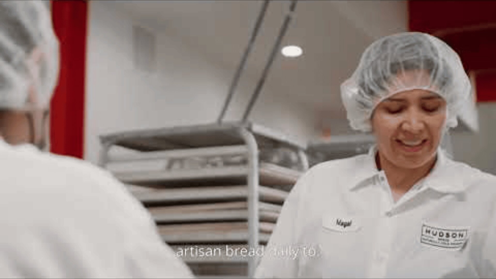A bakery worker in a white uniform and hair net smiles while working, with trays stacked in the background. The workers name tag reads Magal.