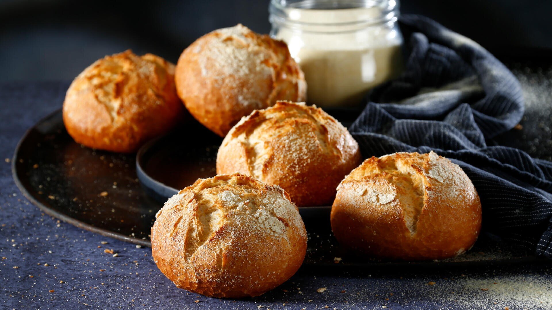 Five round, golden-brown bread rolls dusted with flour are arranged on a dark tray. A glass jar of flour and a dark cloth are in the background. The scene is softly lit and rustic.