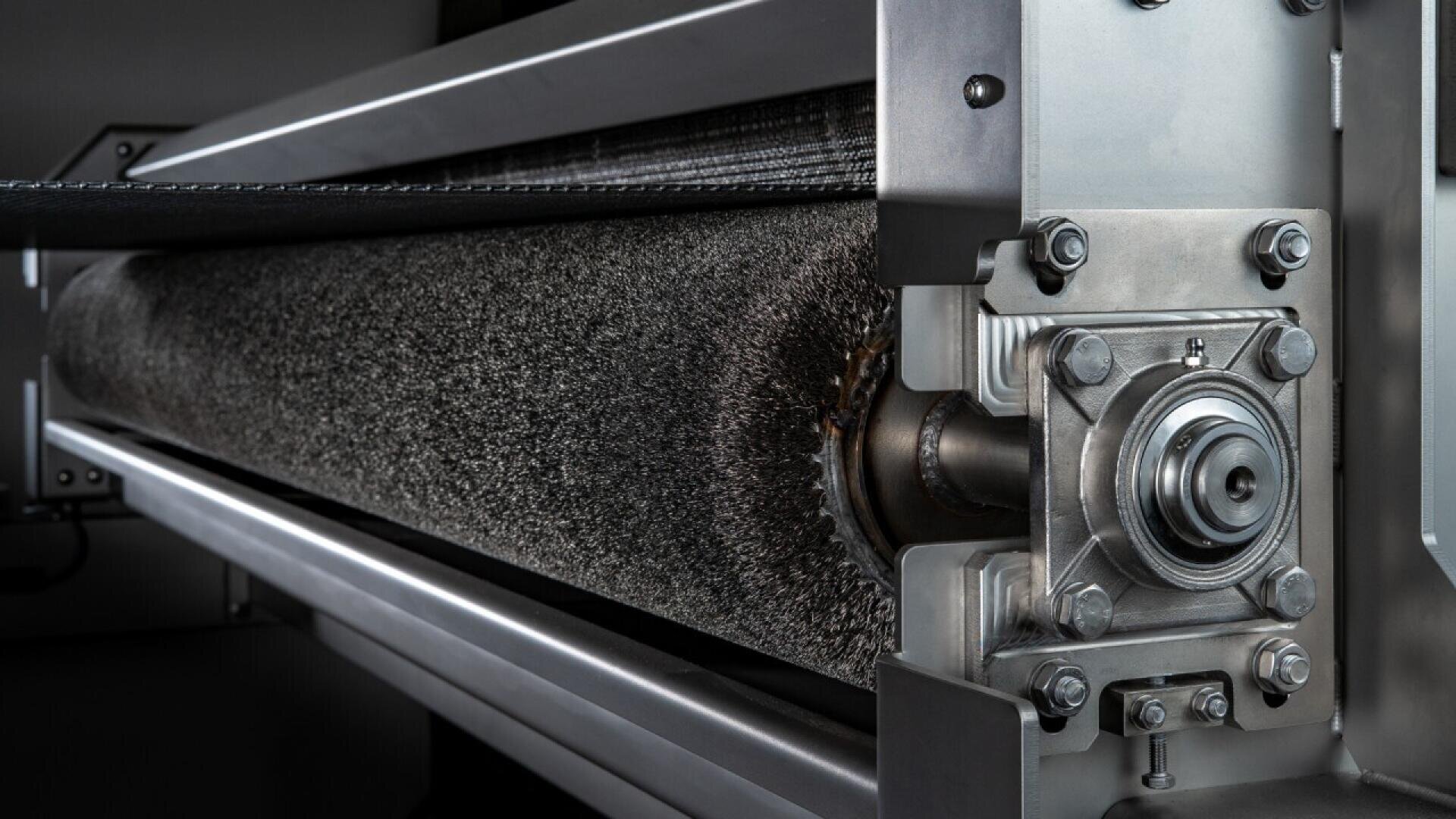 Close-up view of an industrial metal brush roller mechanism, showing detailed metal components, bolts, and textured bristles within a manufacturing or processing machine.