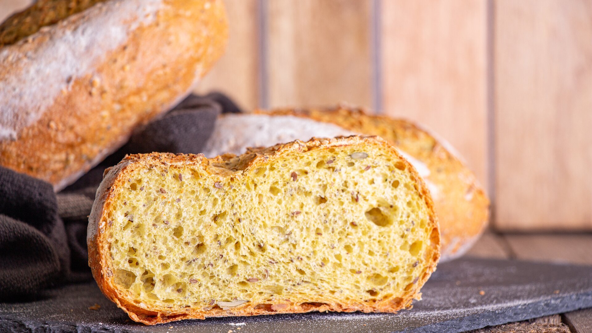 A close-up of a sliced loaf of rustic bread with a golden, airy crumb and textured crust, displayed on a dark surface with more bread and a wooden background in soft focus.