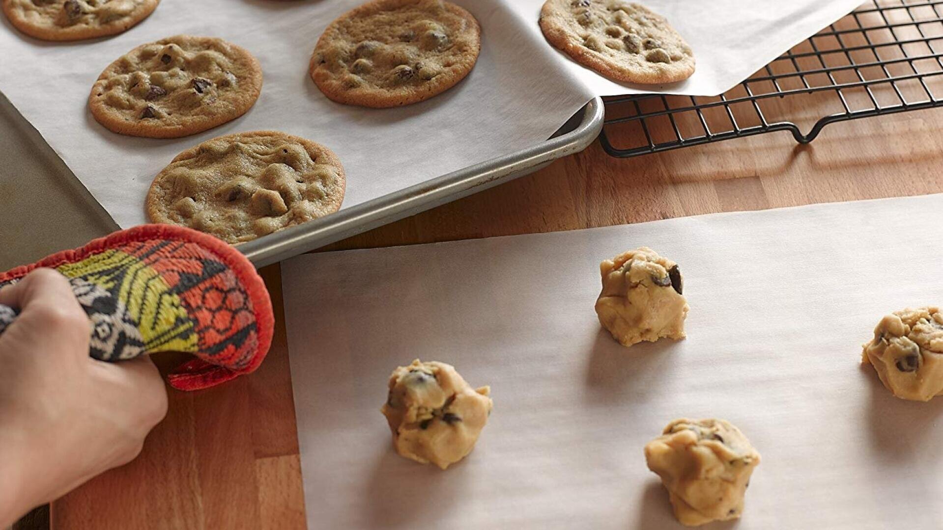 A hand with an oven mitt removes a baking tray of chocolate chip cookies from the oven while unbaked cookie dough balls sit on parchment paper nearby. A cooling rack holds more baked cookies.