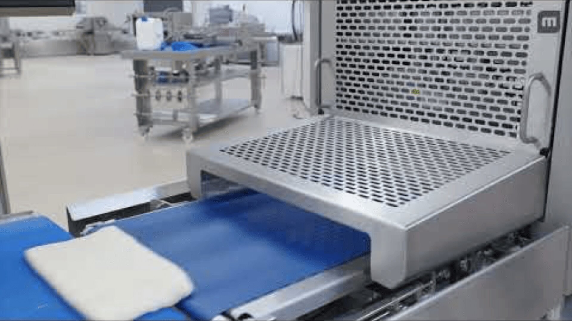 A piece of dough moves on a blue conveyor belt toward a metal machine with a perforated tray in a food processing facility.