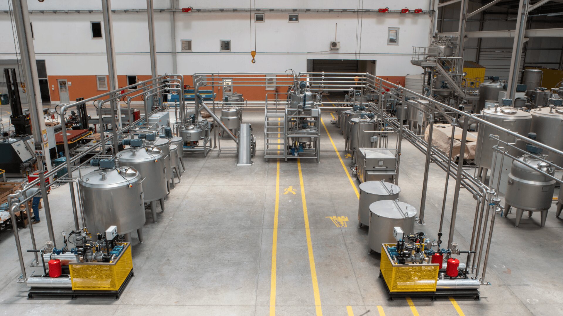 Weitwinkelansicht einer modernen Industriehalle mit großen Edelstahltanks, Maschinen, Rohren und einem sauberen, organisierten Arbeitsbereich mit gelben Sicherheitslinien auf dem Boden.