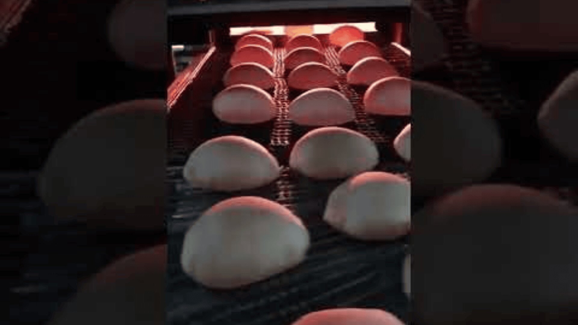 Rows of round dough balls on a conveyor belt move toward an oven, likely in a commercial bakery setting. Warm lighting from above highlights the dough as it bakes.