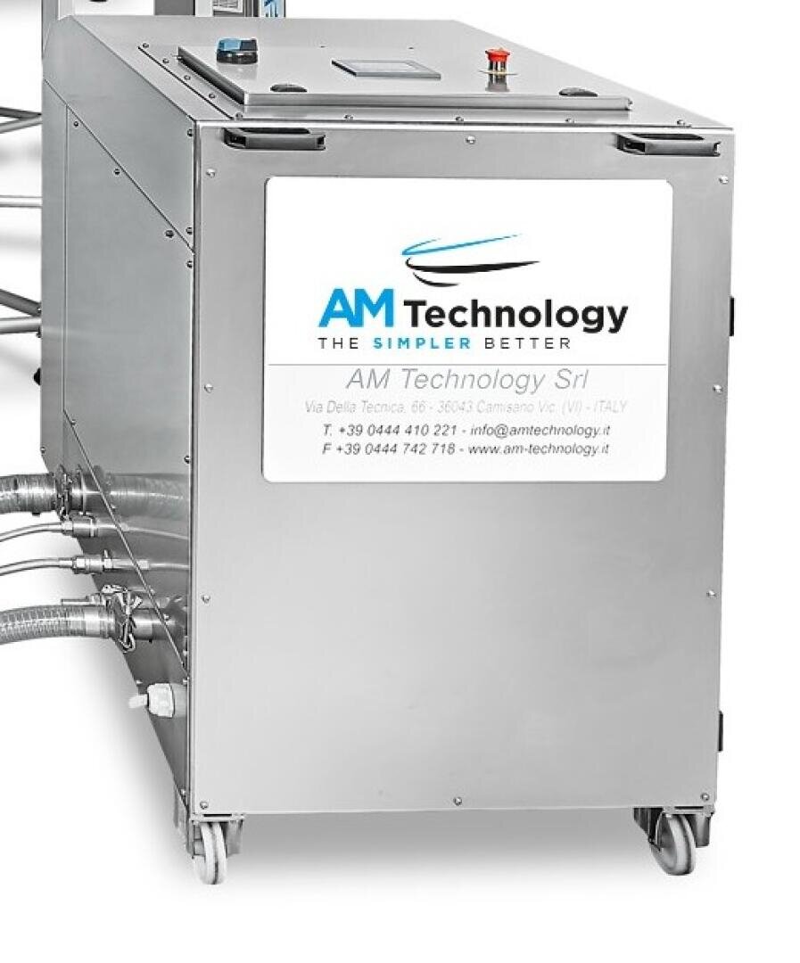 A stainless steel industrial machine on wheels with a sign reading AM Technology and company contact information displayed on the front panel. Several cables and pipes are connected to the side.