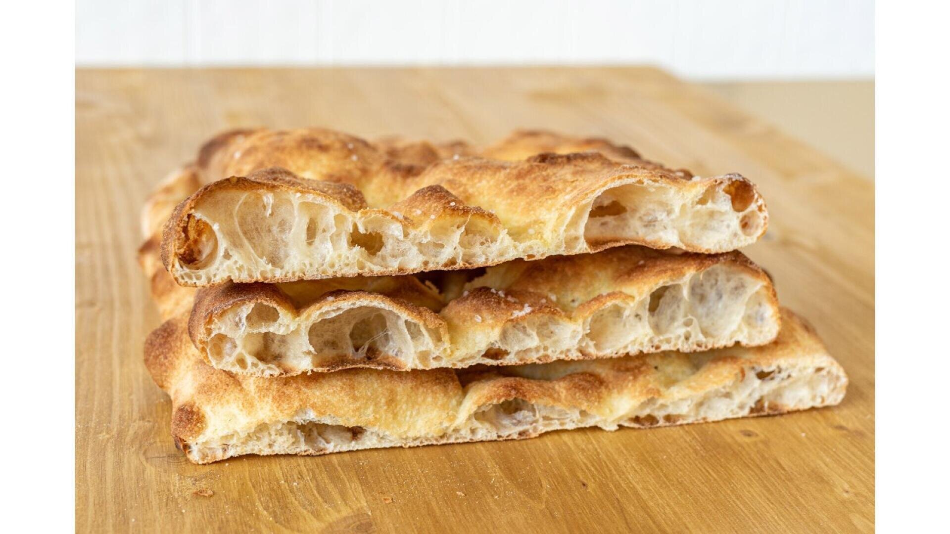 Drei Scheiben luftiges, goldbraunes Focaccia-Brot sind auf einer Holzunterlage gestapelt und zeigen ihre knusprige Kruste und ihre offene, blasige Krumenstruktur.