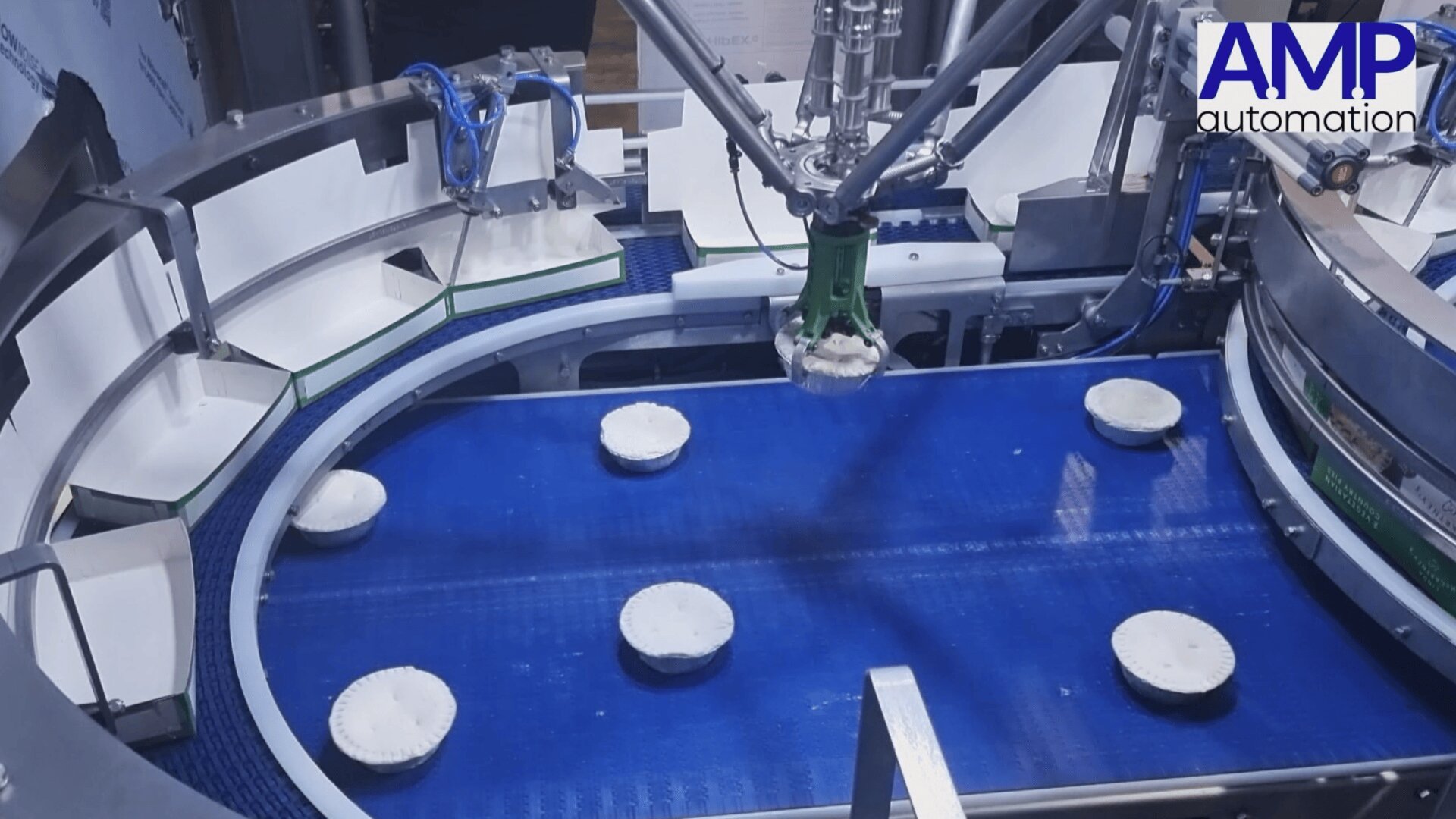 A robotic arm with a green gripper picks up round white objects from a blue conveyor belt in an automated food processing facility. The AMP automation logo is visible in the top right corner.