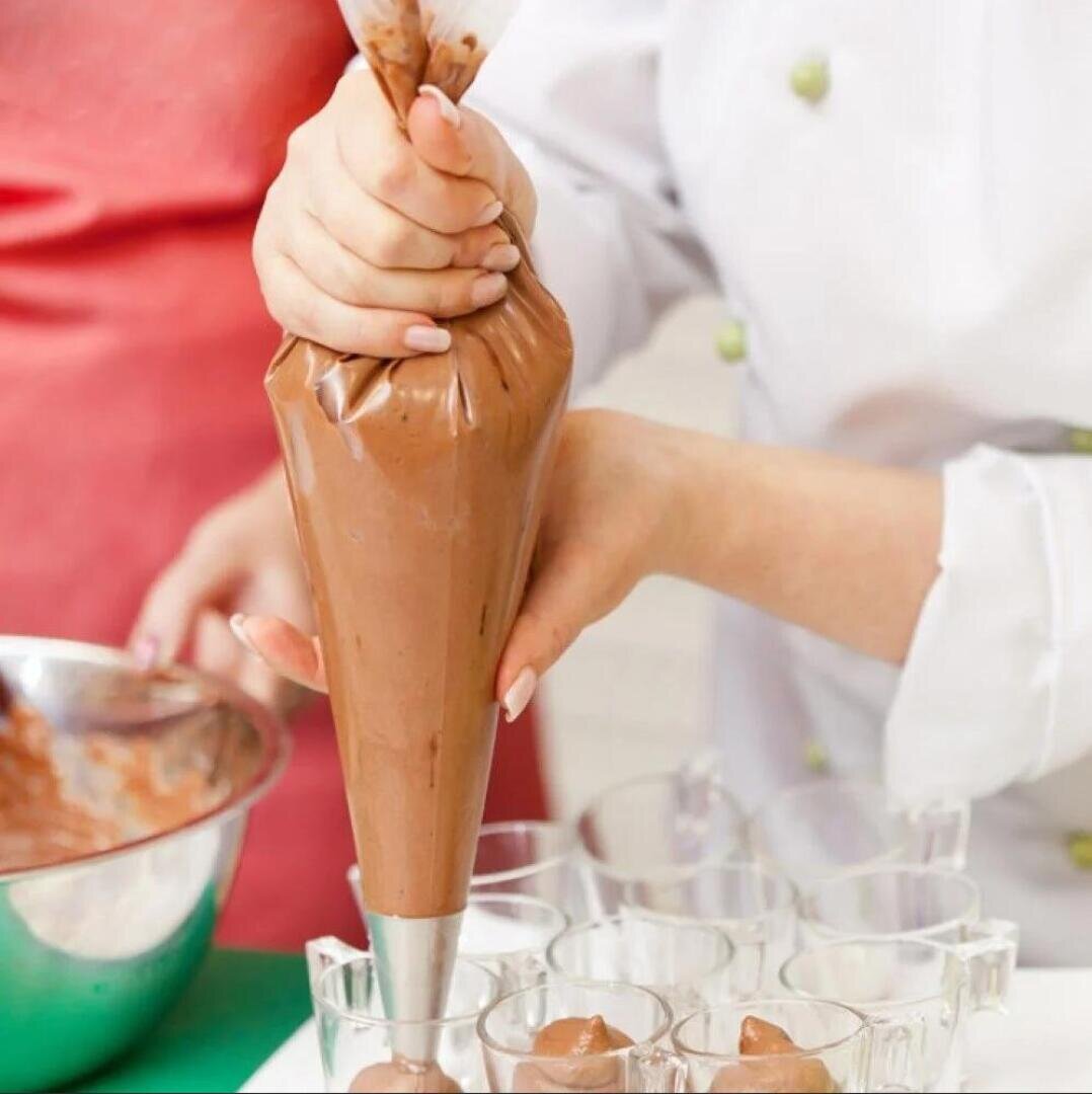 A person uses a piping bag filled with chocolate mousse to fill small glass cups. Another person holds a mixing bowl with more mousse in the background.