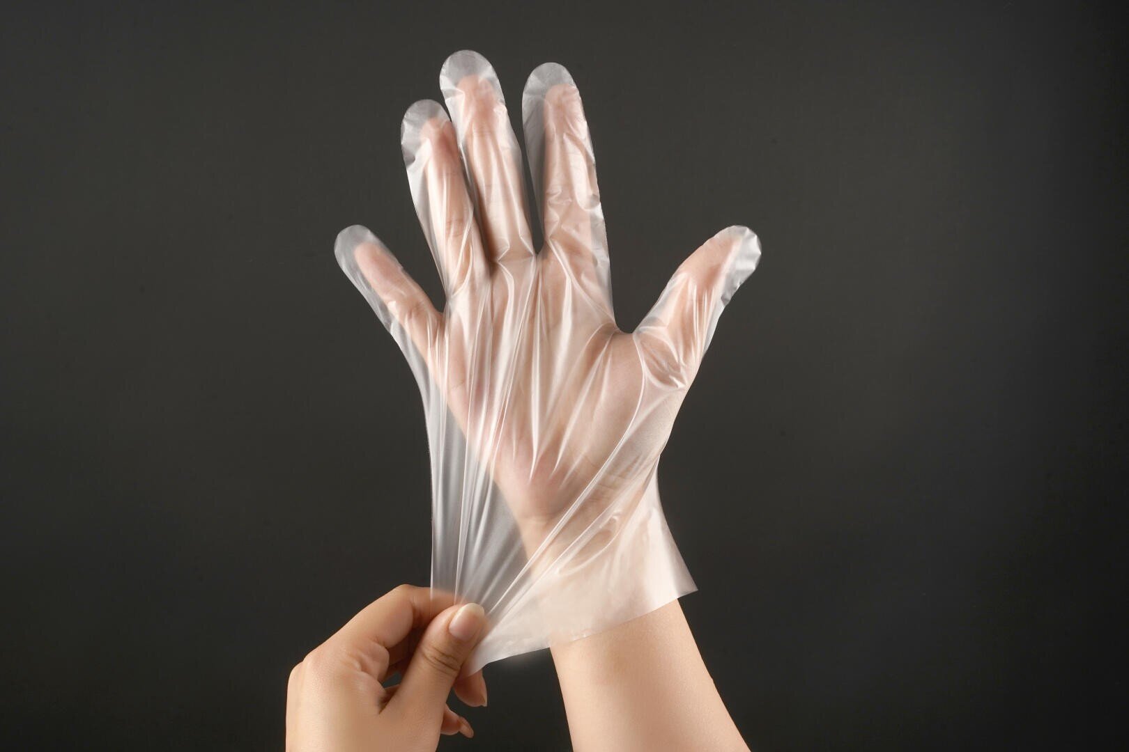 A person puts on a transparent disposable glove against a plain black background, using one hand to pull the glove onto the other hand.