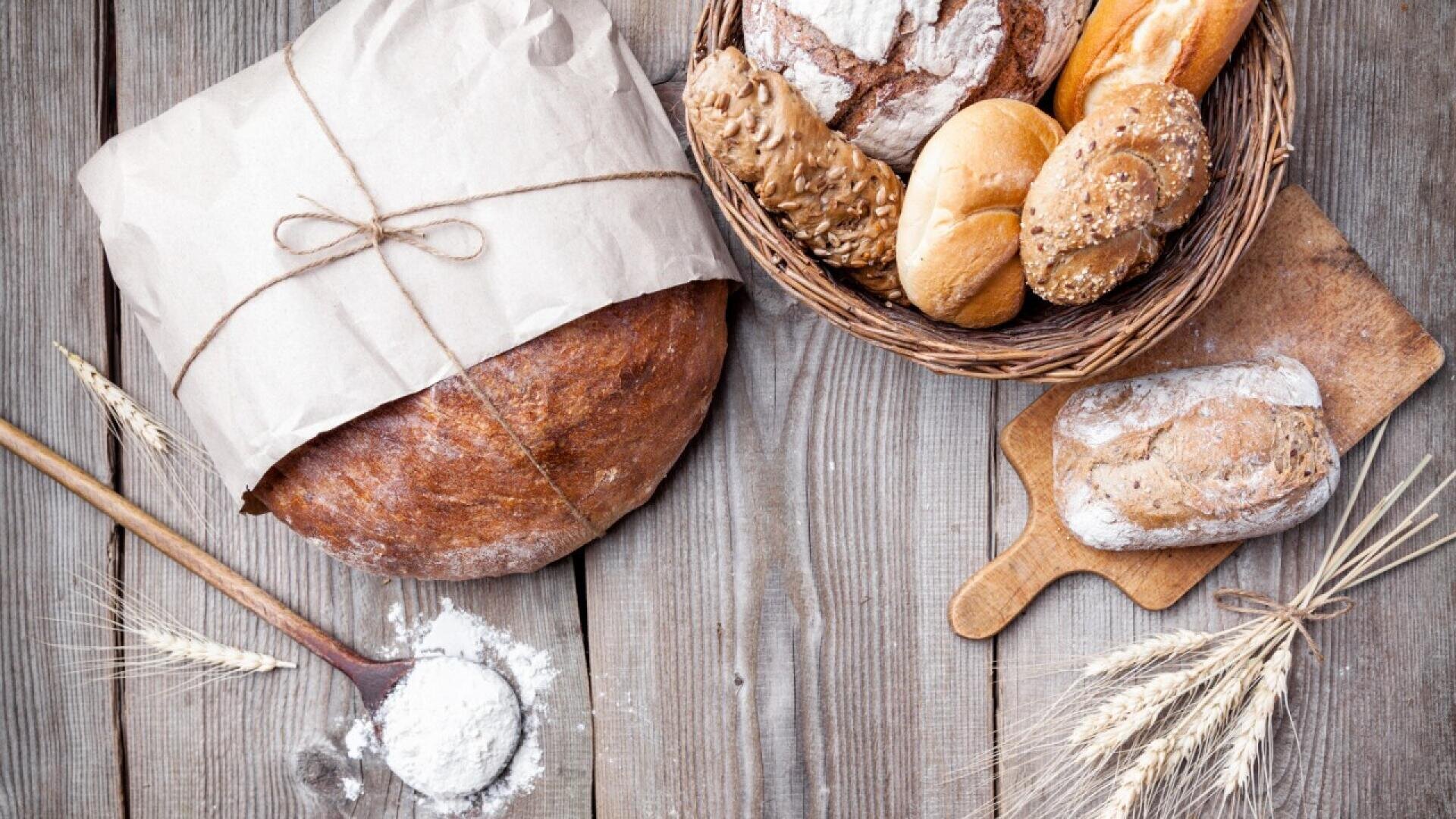 Assorted bread loaves and rolls in a basket and on a wooden board, with a large round loaf wrapped in paper, a wooden spoon with flour, and wheat stalks on a rustic wooden table.