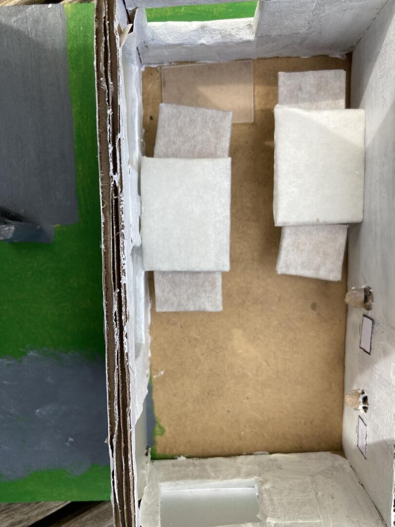 Draufsicht auf ein kleines Modellzimmer mit zwei parallel zueinander aufgestellten Betten aus weißem Stoff. Das Zimmer hat weiße und grüne Wände, einen braunen Boden und kleine Papierfenster.