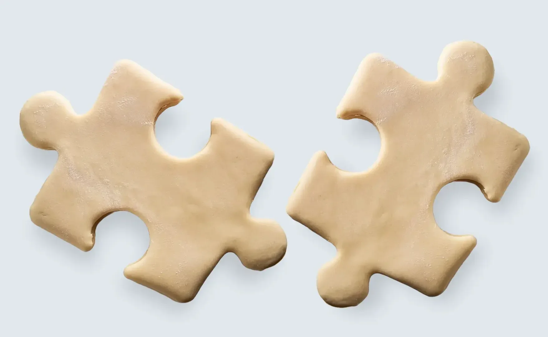 Two light tan puzzle pieces made of dough are placed on a light background, positioned close together but not connected.
