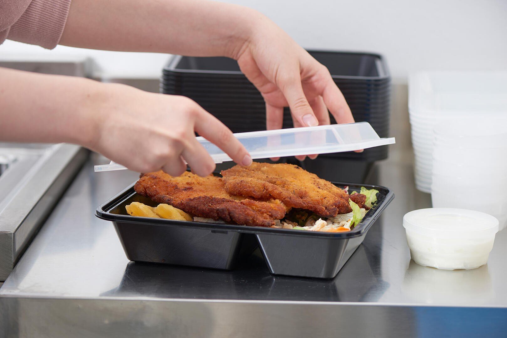 Eine Person legt einen Plastikdeckel auf einen schwarzen Behälter, der mit paniertem Brathähnchen, Kartoffelecken und Salat gefüllt ist. Weiße Soße in einem kleinen Becher steht daneben auf einer Arbeitsplatte.