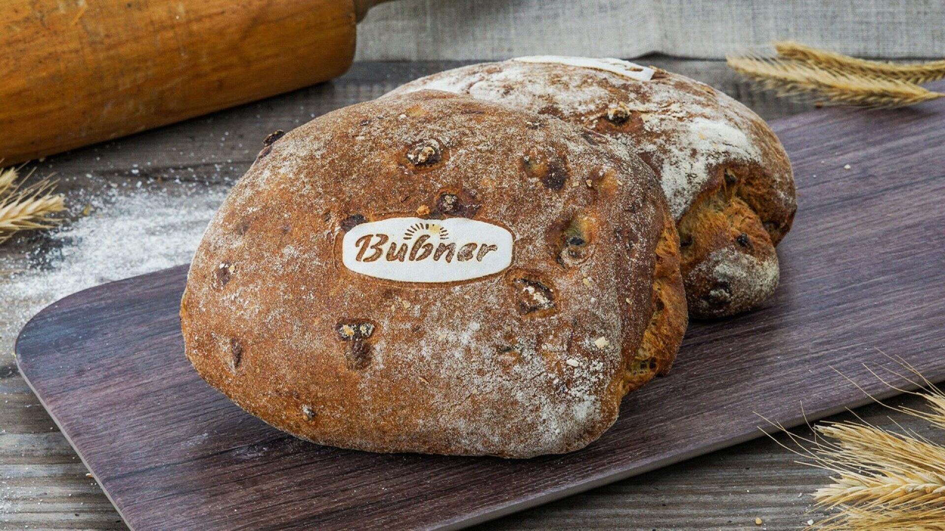 Two rustic loaves of bread with Bubner labels rest on a wooden board, surrounded by wheat stalks and a rolling pin, with some flour sprinkled on the surface.