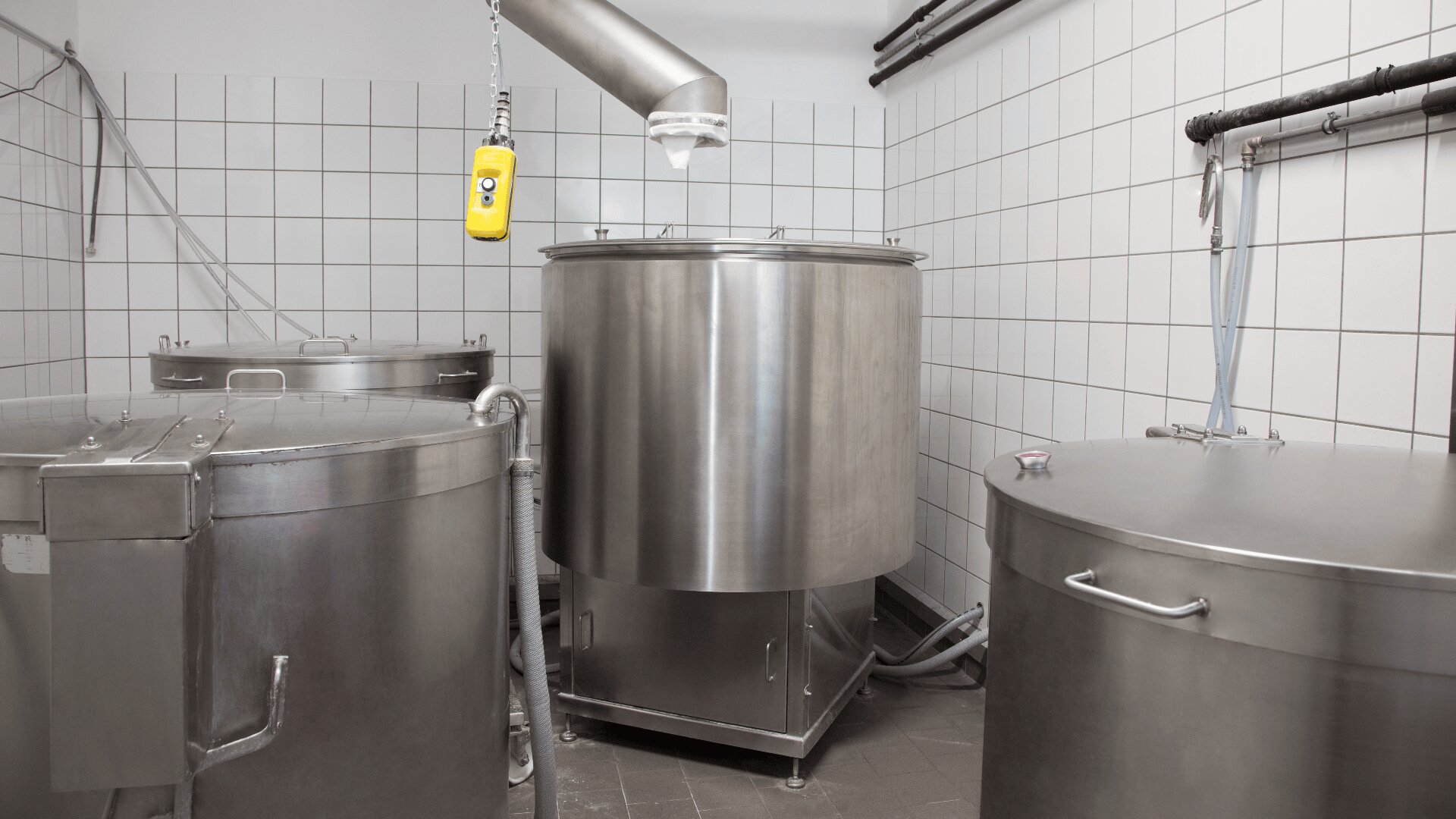 In einem weiß gefliesten Raum stehen drei große Industrietanks aus Edelstahl mit Deckel. Von der Decke hängen Rohre und eine gelbe Steuerzentrale, die wahrscheinlich zur Lebensmittel- oder Getränkeverarbeitung verwendet wird.