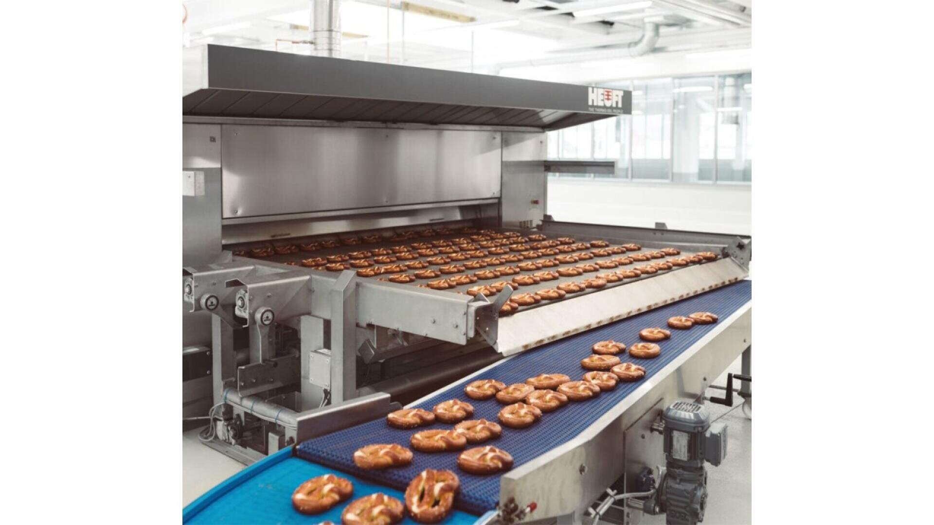Pretzels move along a conveyor belt in a large industrial bakery, emerging from an automated oven in a clean, modern facility.