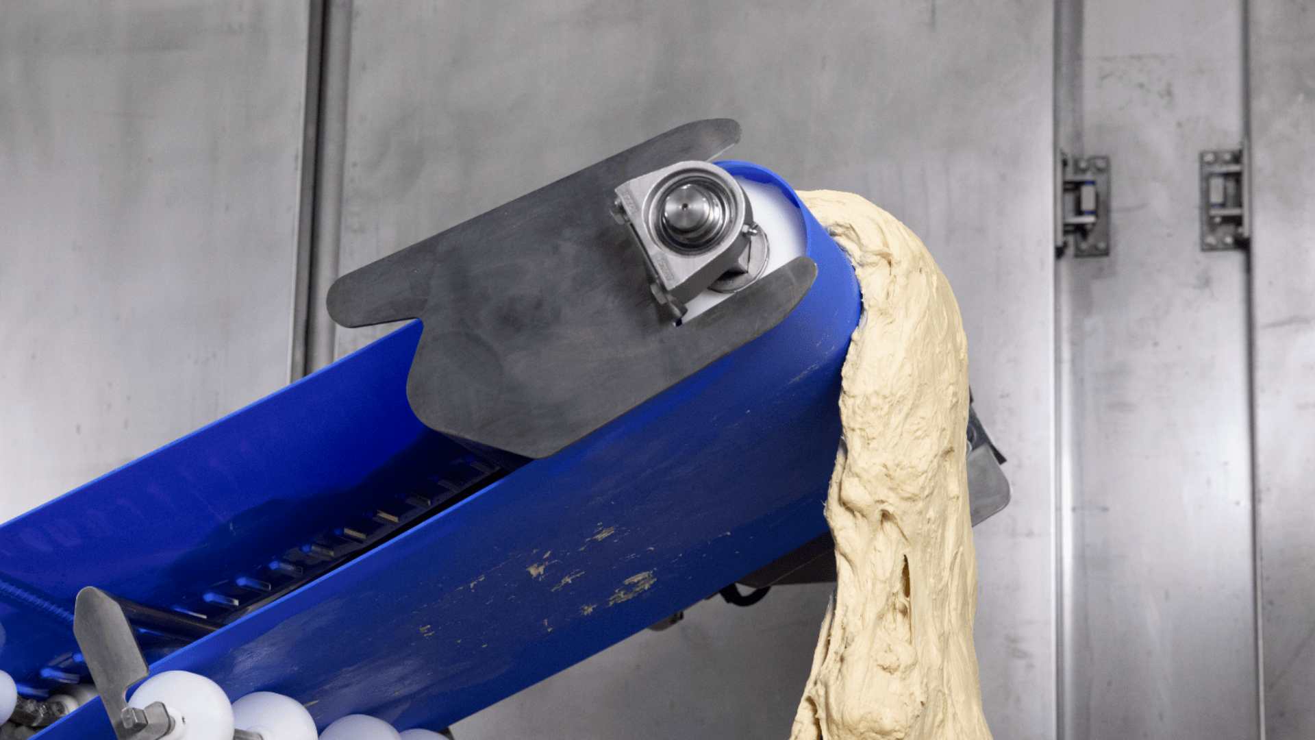 Dough being transported upwards on a blue conveyor belt inside a processing facility with metal walls in the background.