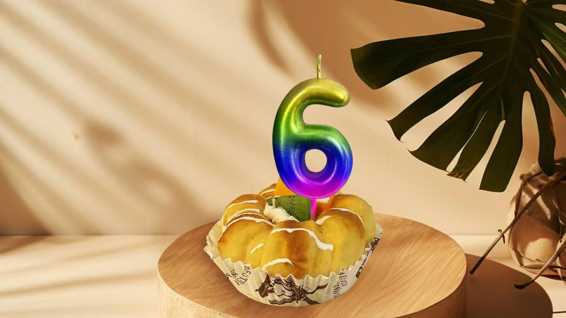 A pastry with icing is topped with a colorful number 6 candle, placed on a wooden surface. Sunlight and shadows from a large green plant leaf are visible in the background.