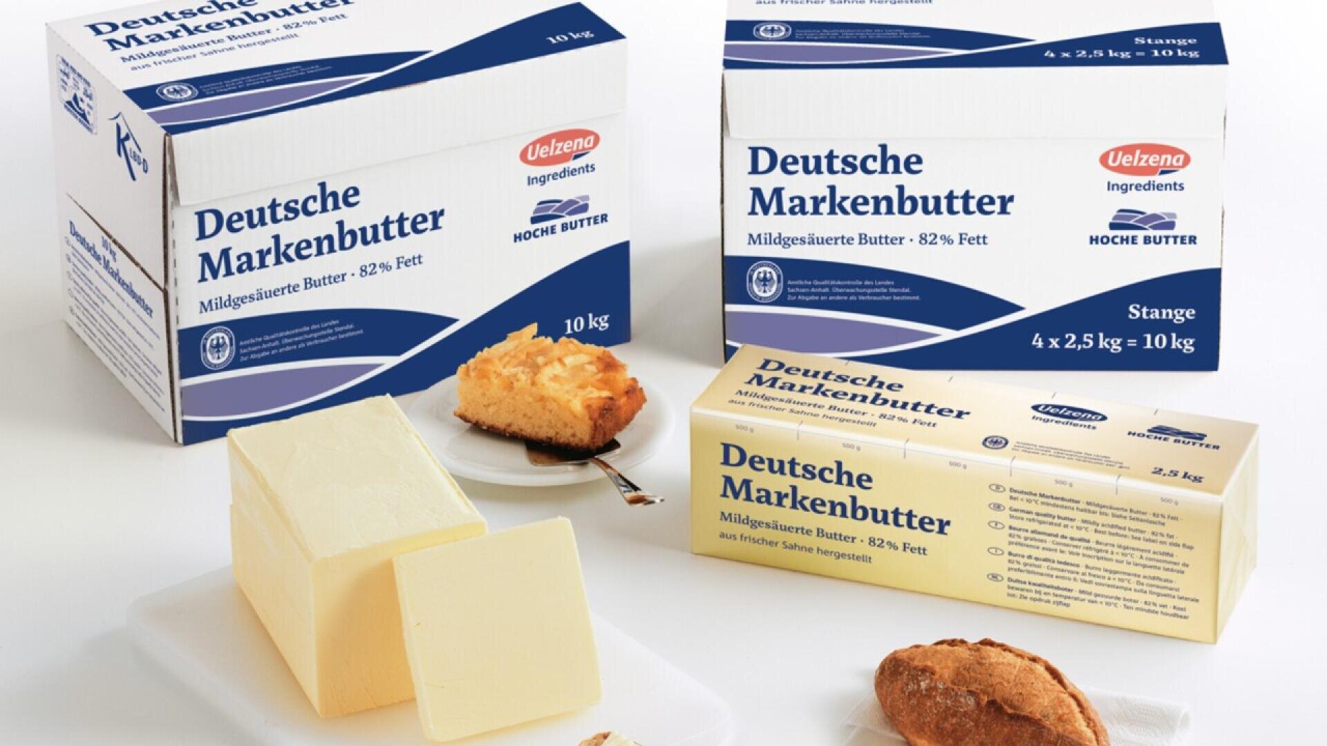 Boxes and a stick of Deutsche Markenbutter butter are displayed on a table with slices of bread, a piece of cake, and portions of butter. The packaging is white and blue with German text.