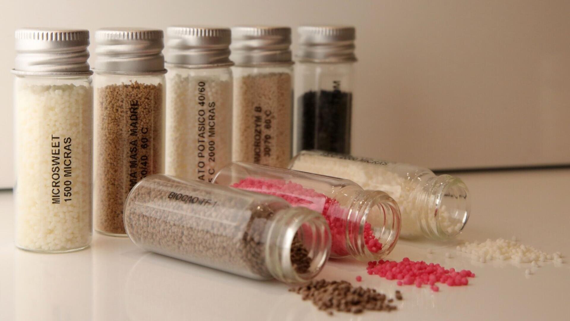 Sechs kleine Fläschchen gefüllt mit verschiedenfarbigen Körnchen, einige aufrecht und einige auf der Seite liegend, wobei eine Mischung aus weißen, braunen, rosa und schwarzen Pellets auf eine reflektierende Oberfläche herausfällt.