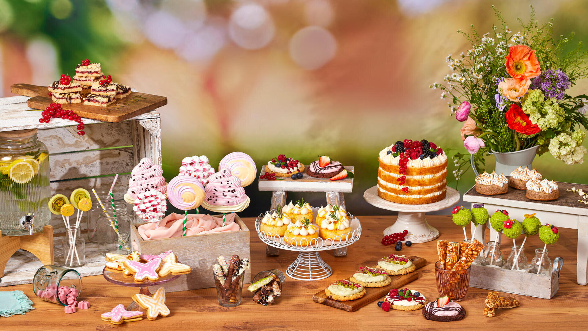 Ein farbenfroher Desserttisch im Freien mit Kuchen, Cupcakes, Baisers, Keksen, Cake Pops, Bonbons und einem Limonadenspender. Auf der rechten Seite des Holztisches steht ein Blumenstrauß.