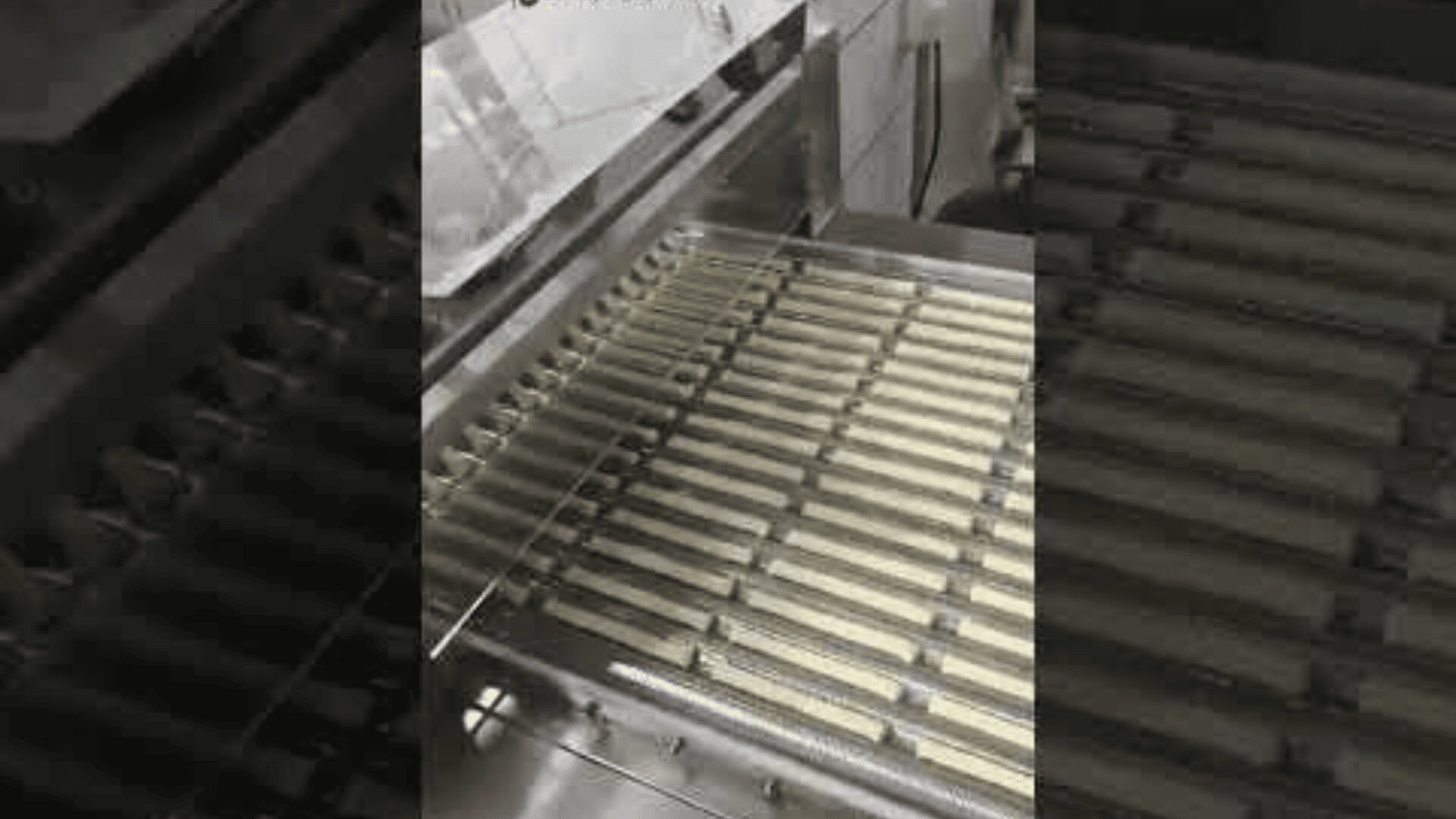 A close-up of a machine in a factory, with rows of long, rectangular biscuits or cookies being processed on a metal conveyor.