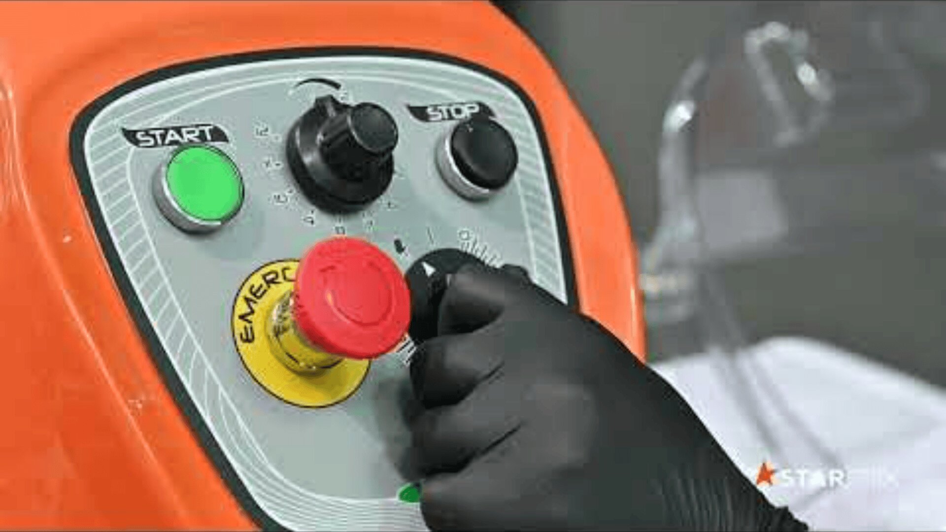 A person wearing a black glove presses a red emergency stop button on an orange machine control panel with start, stop, and dial switches.
