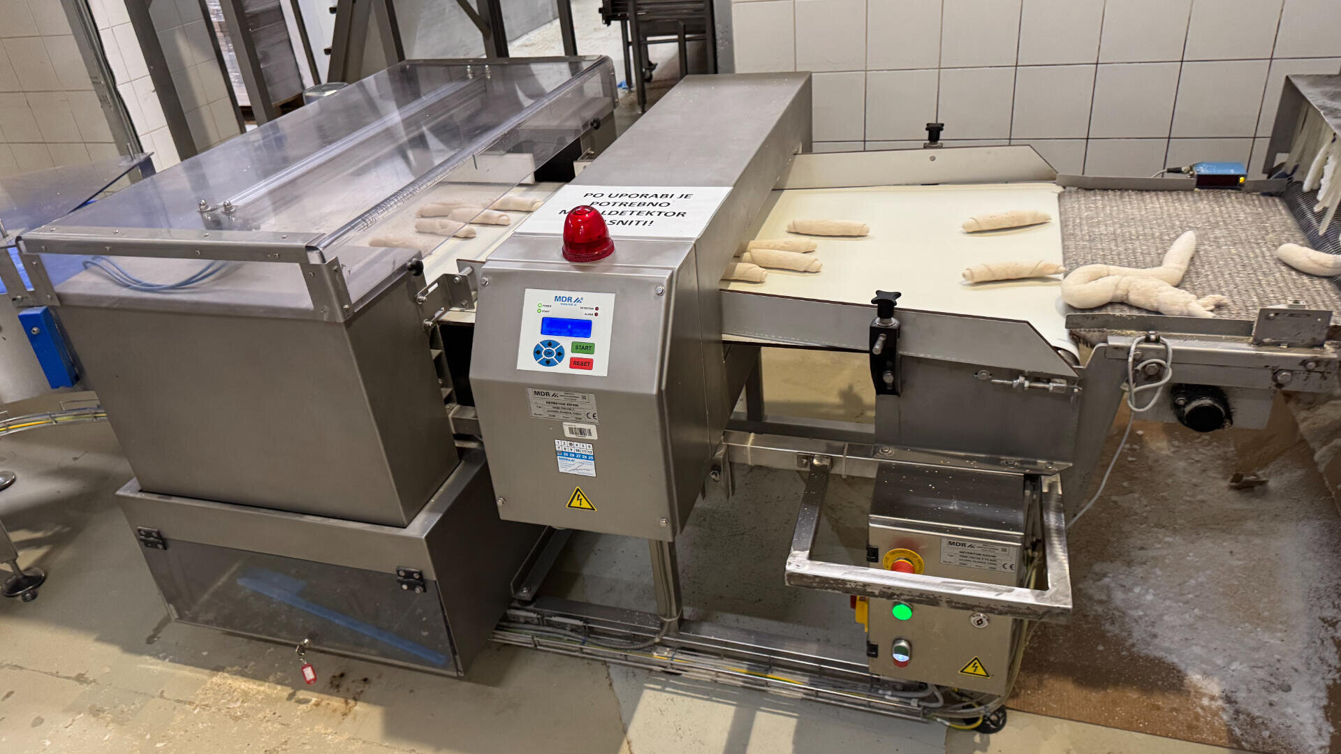 A commercial bakery machine processes and shapes dough pieces on a conveyor belt, with control panels and safety labels visible on the stainless steel equipment.