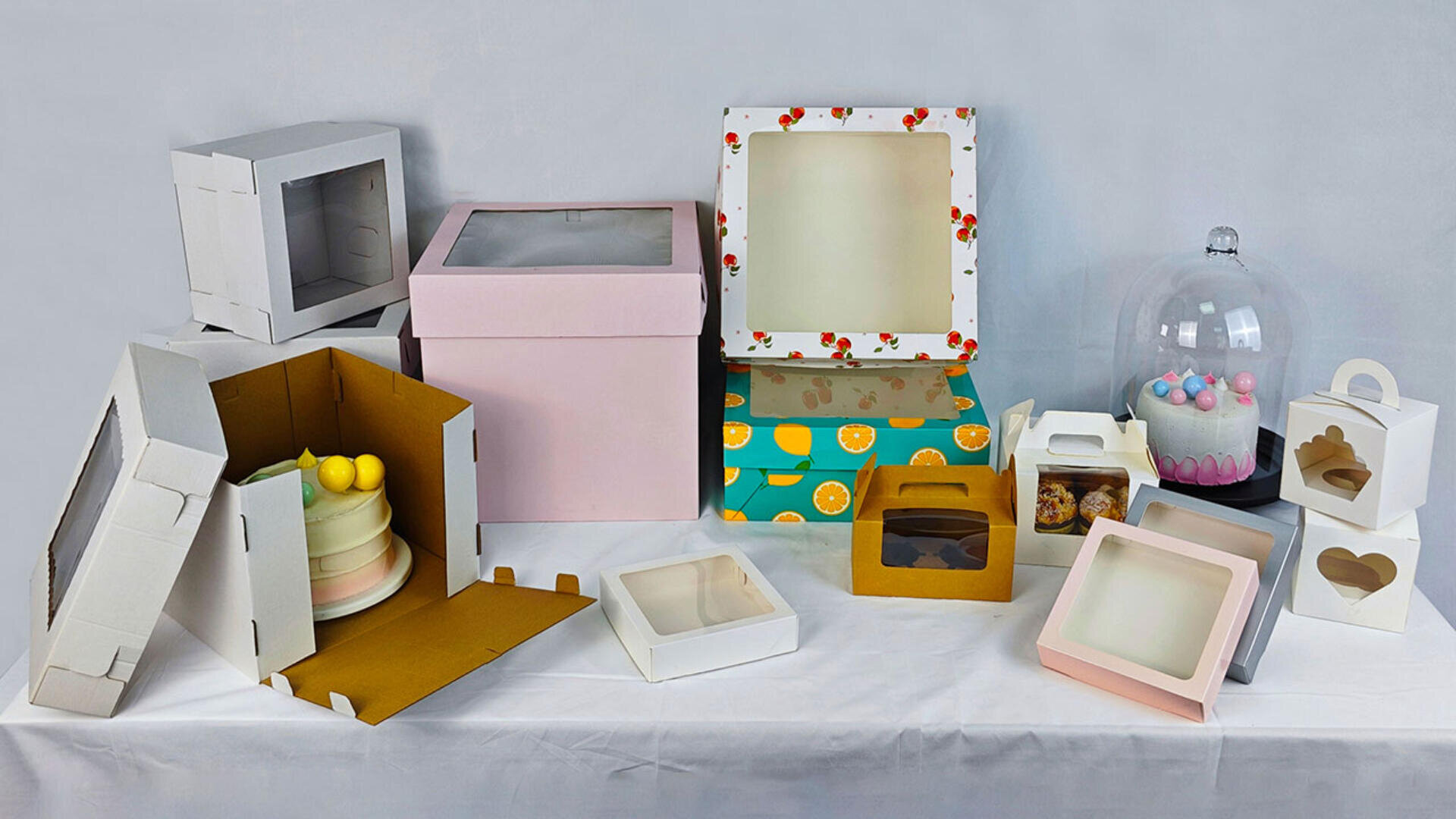 A variety of decorative and plain cake and pastry boxes in different shapes, sizes, and colors are displayed on a white table, along with a glass dome covering cupcakes and a cake in a partially open box.