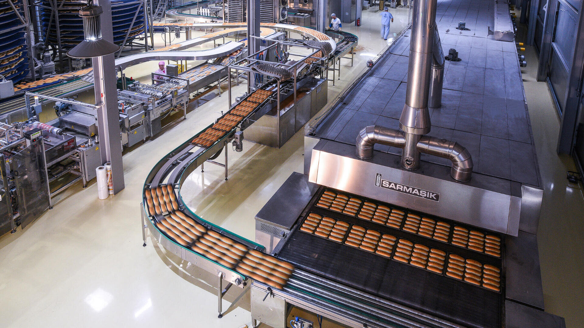 Conveyor belts in a large industrial bakery transport rows of freshly baked bread loaves through machinery, with workers in protective clothing visible in the background.