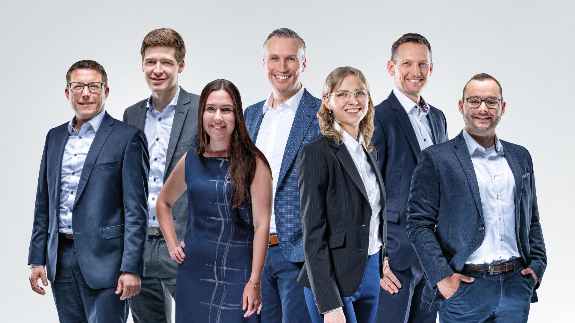 Seven professionally dressed adults, five men and two women, stand in a row against a light gray background, smiling and posing confidently for a group photo.