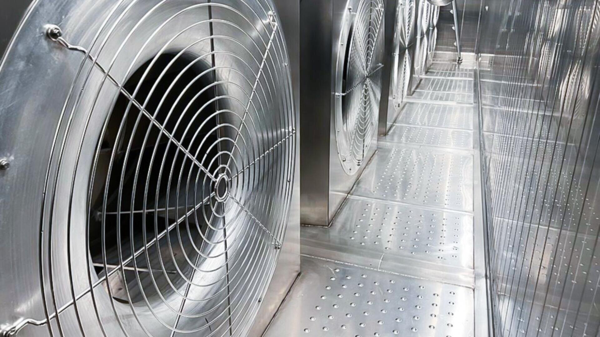 Nahaufnahme großer industrieller Metallventilatoren und perforierter Metallböden in einem Lüftungs- oder Kühlsystem aus Edelstahl. Die glänzenden Oberflächen reflektieren das Licht und betonen die saubere, moderne Umgebung.