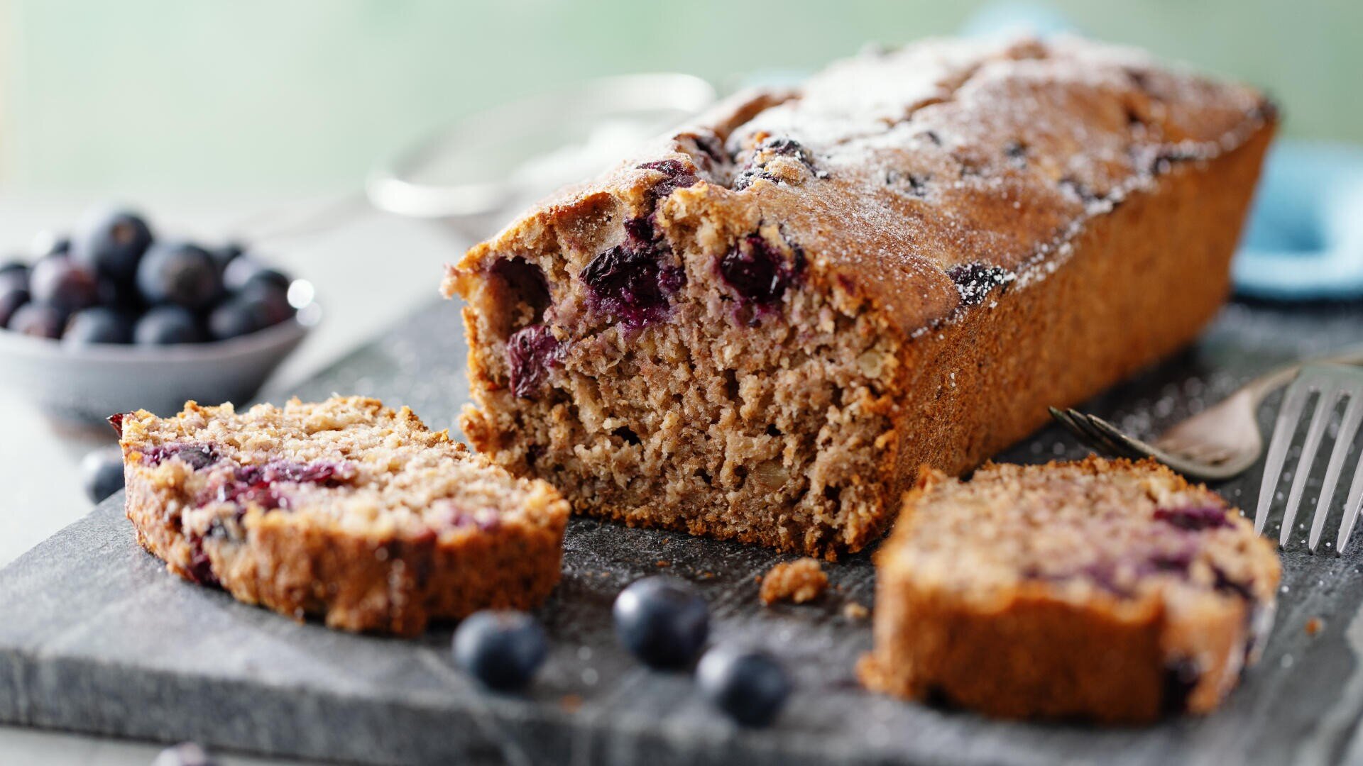 A loaf of blueberry bread with a slice cut, showing a moist interior with visible blueberries, sits on a dark cutting board. Loose blueberries and a fork are nearby, with a bowl of blueberries in the background.