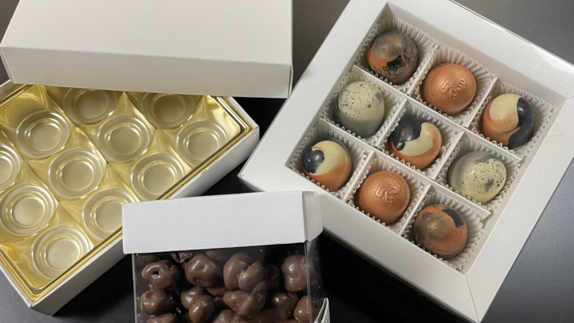 Three white boxes of chocolates: one empty with a gold tray, one with nine assorted round chocolates in white paper cups, and one filled with small chocolate-covered treats. All boxes are open and displayed on a dark surface.
