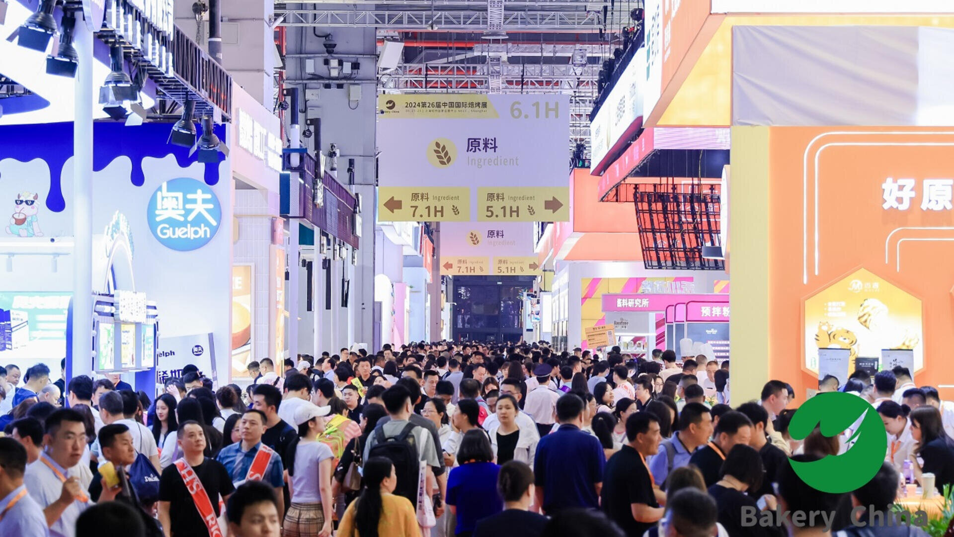 A large crowd of people walks through a brightly lit exhibition hall filled with colorful booths and banners, with signs hanging from the ceiling indicating different areas and sections.