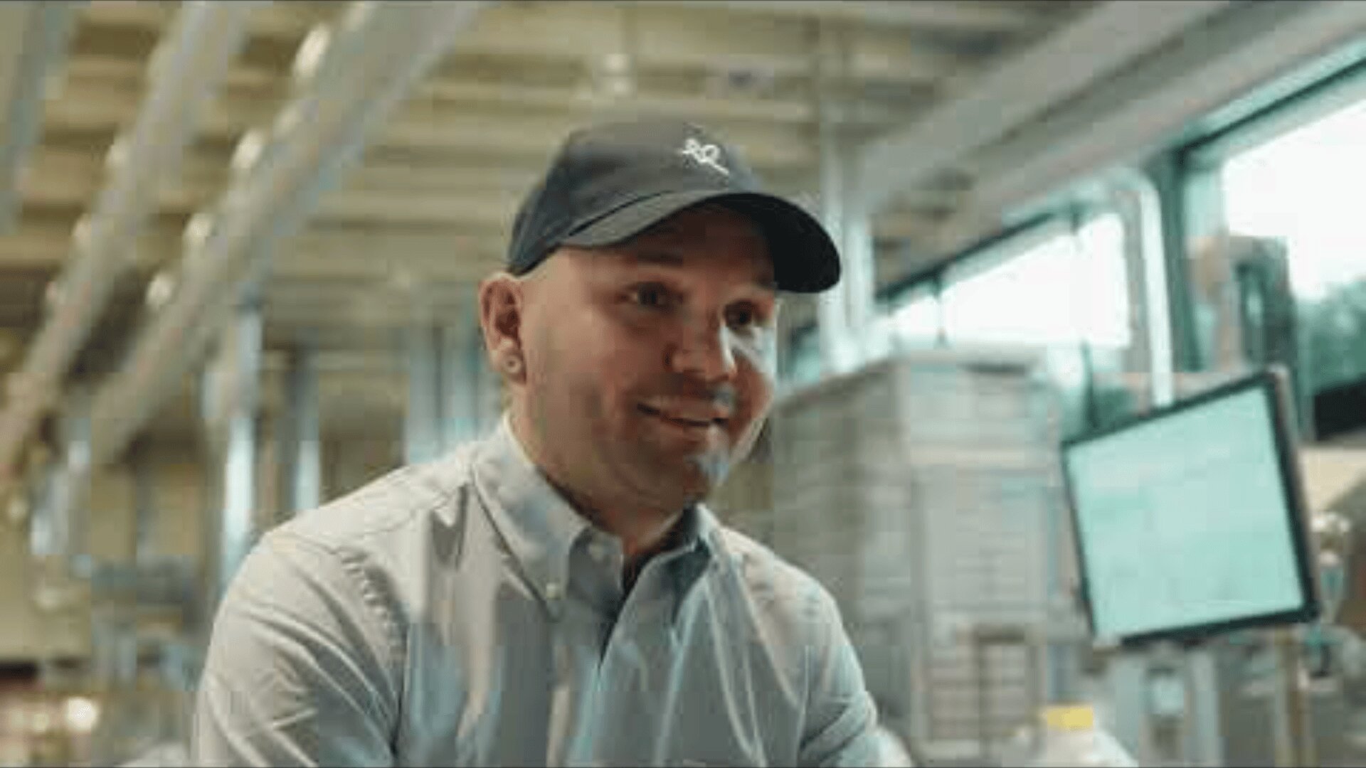 A smiling man wearing a baseball cap and button-up shirt sits indoors, with industrial equipment and a computer monitor visible in the background. The setting appears to be a modern workspace or factory.