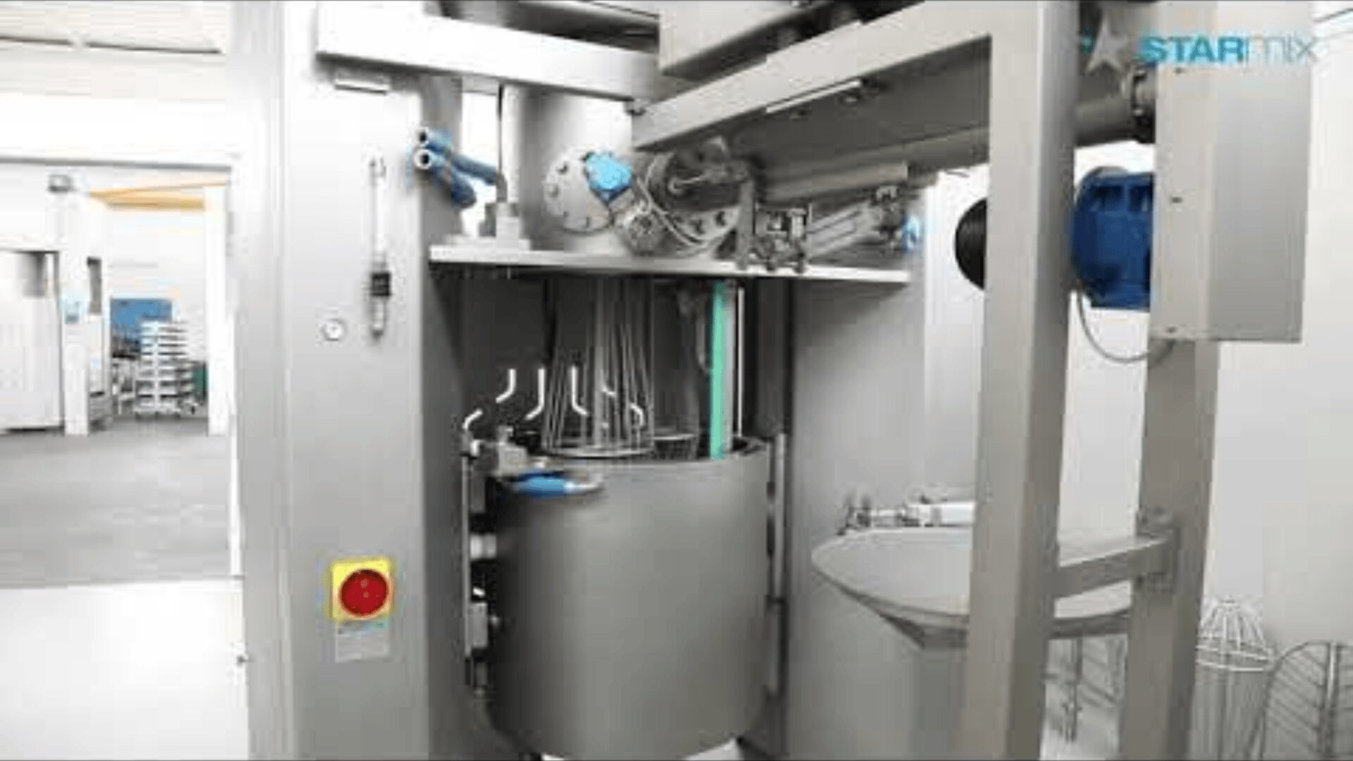 Industrial mixing machine with a large metal bowl, multiple mixing attachments above the bowl, and control buttons on the side, located in a clean, modern facility.