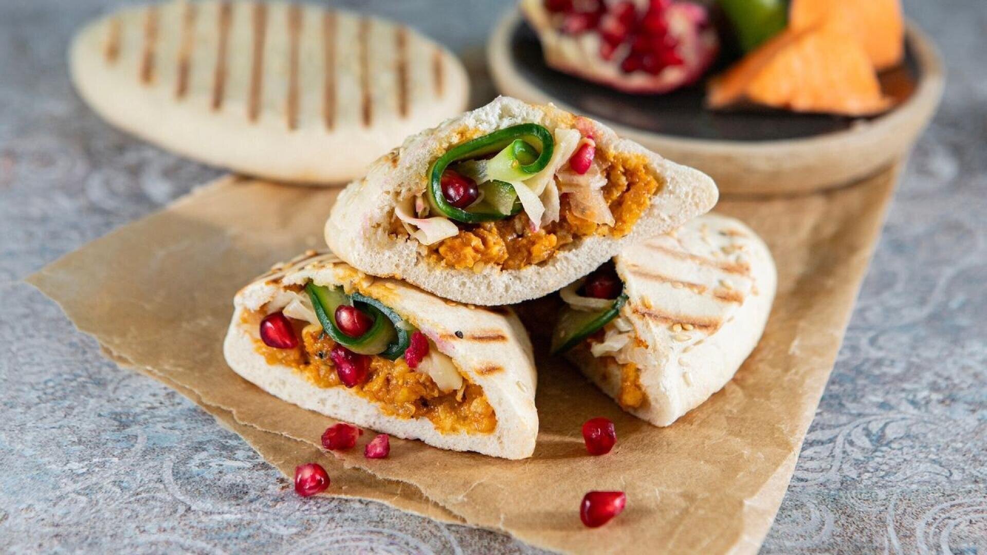 Three pita pockets filled with a mixture of orange spread, sliced vegetables, and topped with pomegranate seeds are stacked on brown parchment paper, with extra pita and pomegranate pieces in the background.