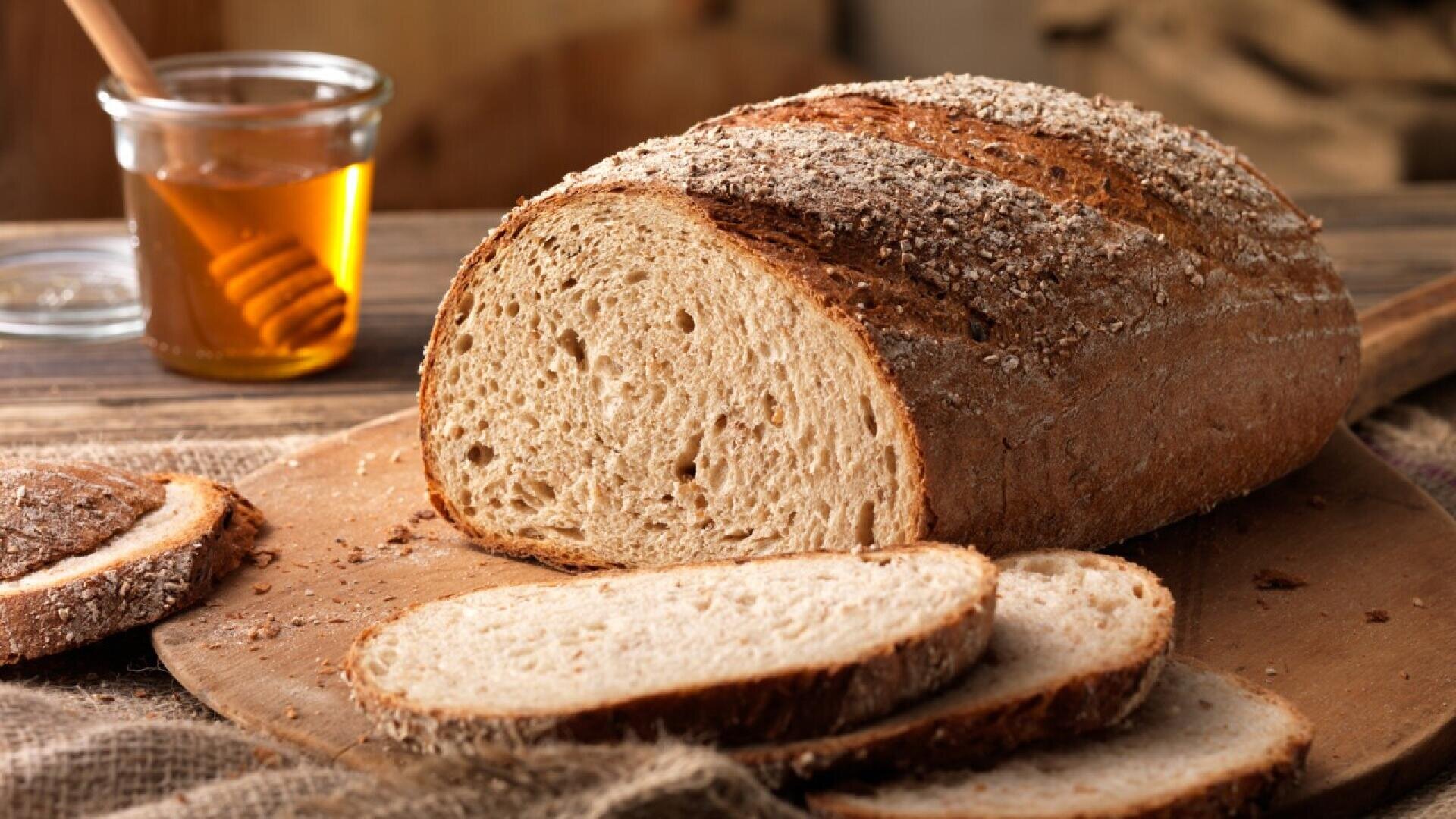 A rustic loaf of sliced whole grain bread sits on a wooden cutting board, with a jar of honey and a honey dipper in the background. The setting exudes a cozy, homemade feel.