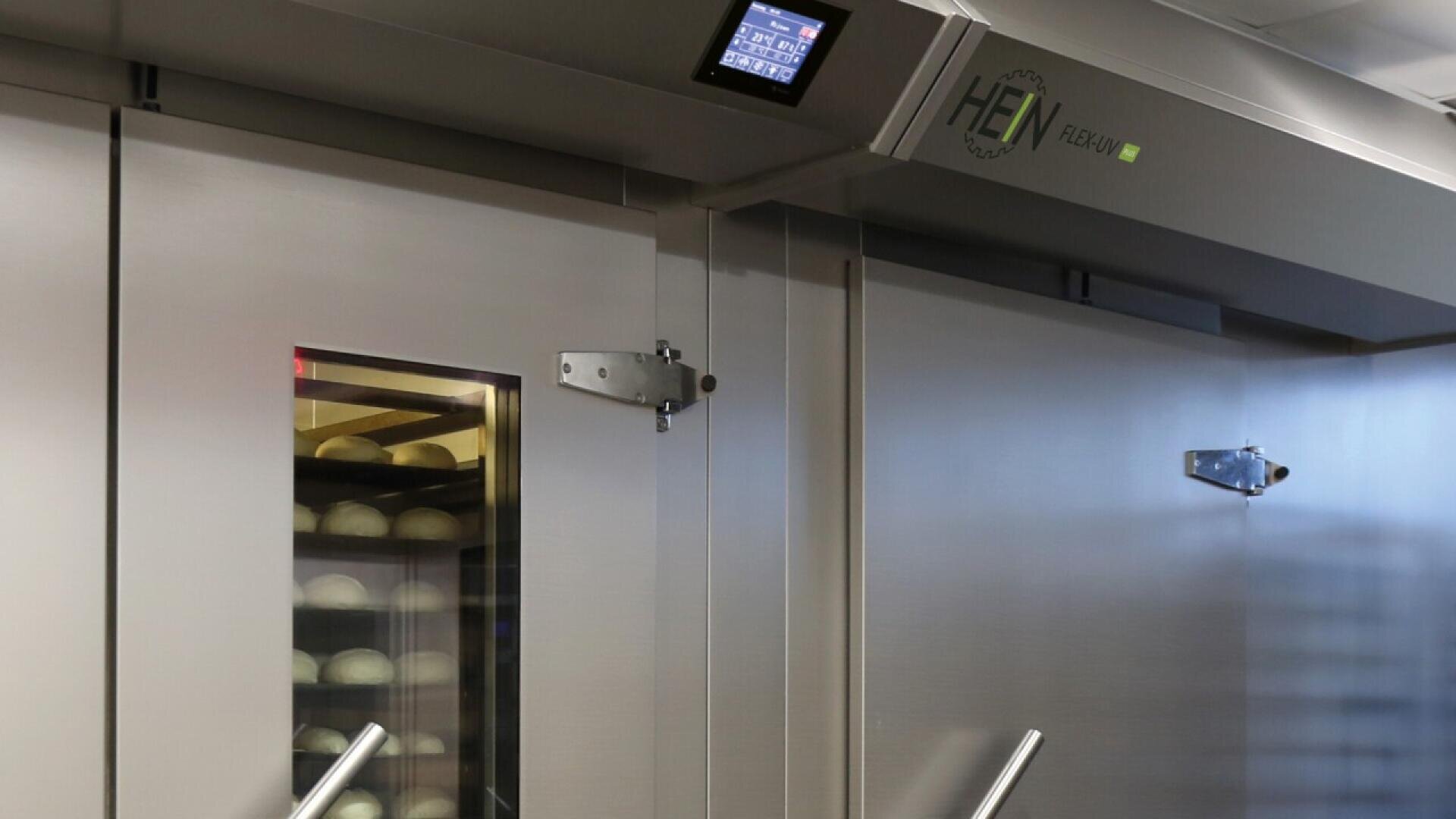 A commercial bakery oven with a glass door showing trays of dough inside, stainless steel handles, and a digital control panel on top.