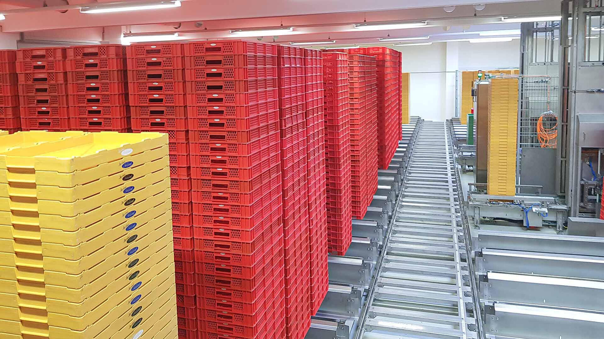 In einer hellen Industriehalle mit weißen Wänden und Deckenbeleuchtung stapeln sich rote und gelbe Plastikschalen neben einem metallenen Fördersystem.