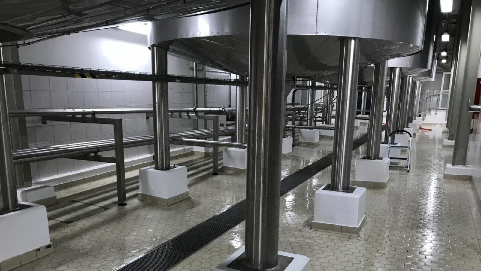 Große Tanks und Rohre aus rostfreiem Stahl in einer sauberen Industrieanlage, möglicherweise einem Lebensmittel- oder Getränkeverarbeitungsbetrieb, mit gefliesten Böden und weiß gekachelten Wänden. Der Bereich erscheint gut beleuchtet und geordnet.