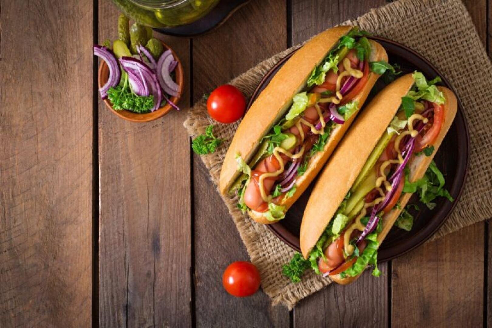 Two loaded sub sandwiches with lettuce, tomato, red onion, pickles, and condiments on a plate, placed on a rustic wooden table with cherry tomatoes and sliced onions nearby.
