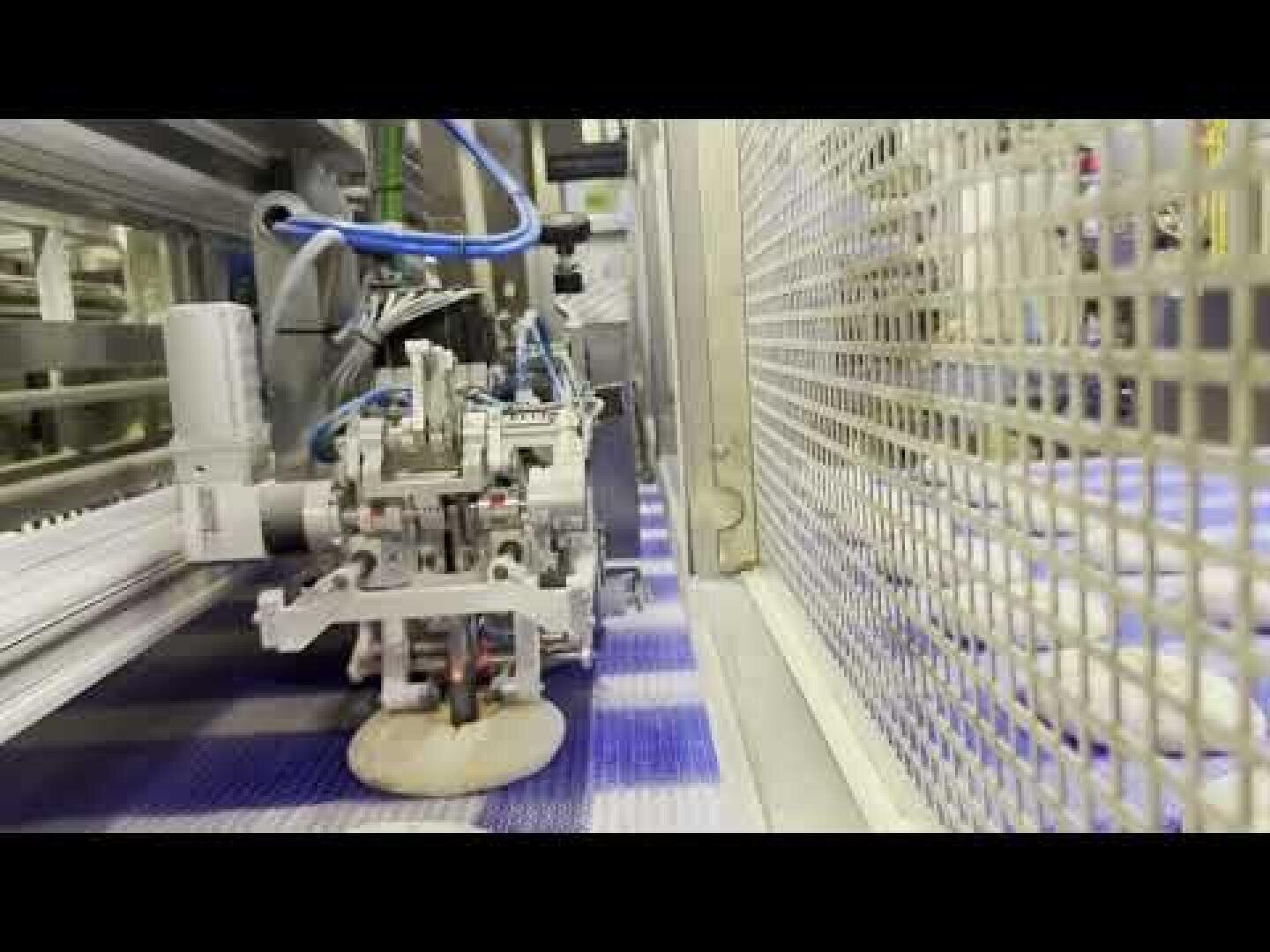A robotic machine places a round piece of dough onto a conveyor belt in a food processing facility, surrounded by metal equipment and fencing.