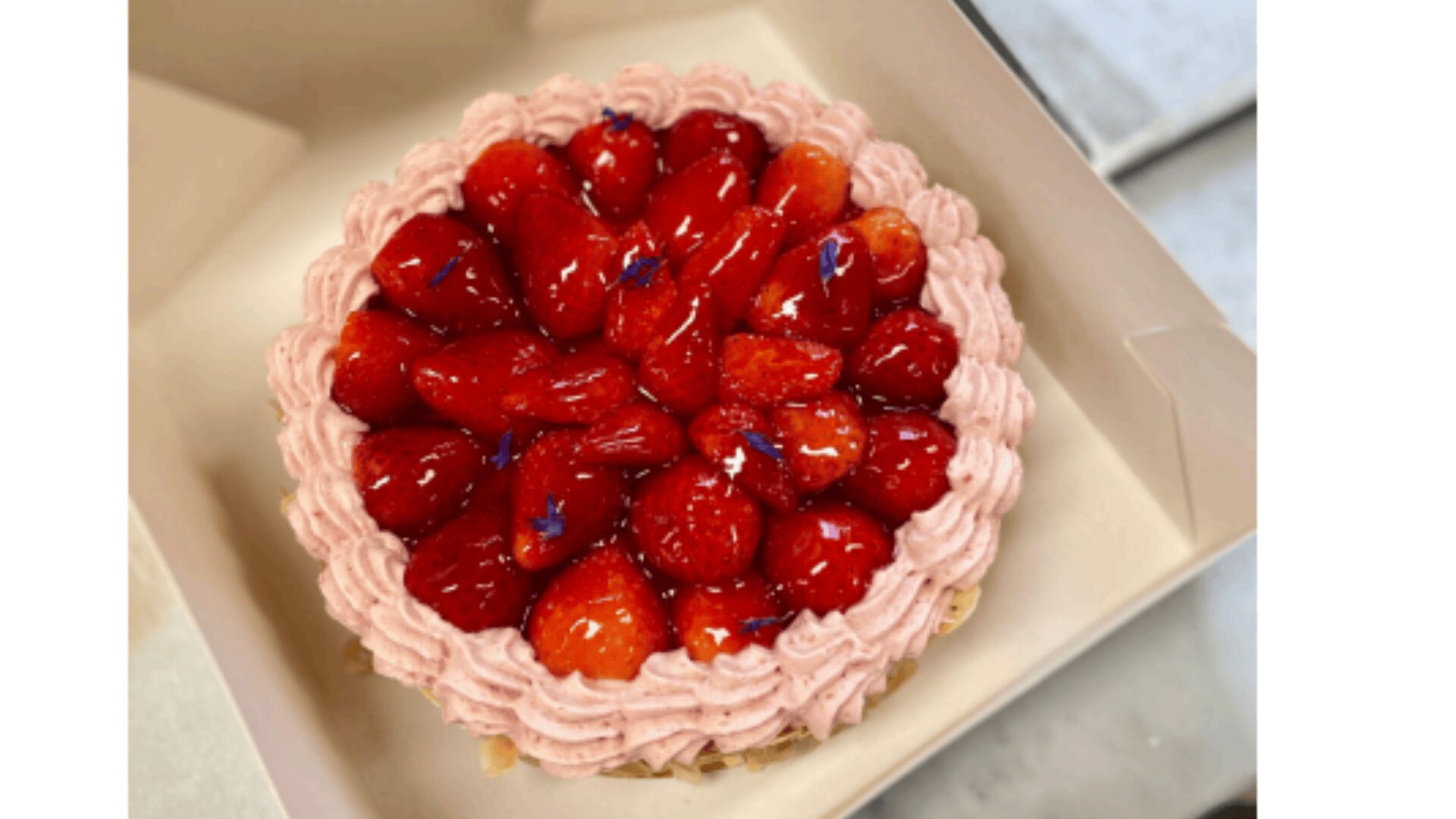 Ein runder Erdbeerkuchen mit rosafarbenem Zuckerguss, gekrönt von ganzen, glänzenden Erdbeeren und kleinen lila Blütenblättern, in einer weißen Schachtel.