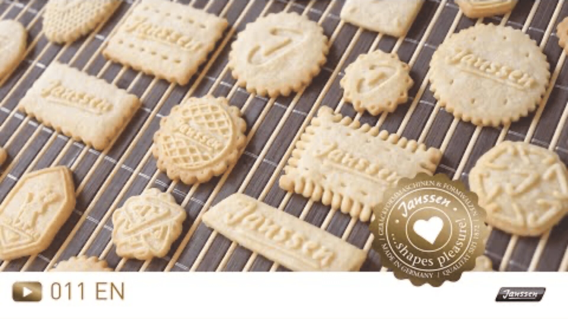 Assorted butter cookies with various shapes, patterns, and the word Janssen embossed on some, are cooling on a wire rack. A gold seal in the corner reads Janssen Shapes Plaisir!.
